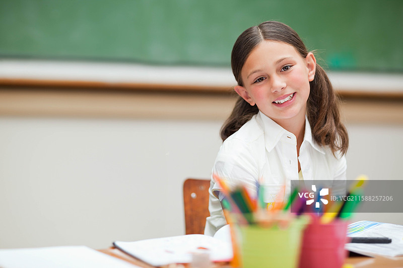 微笑的女学生坐在教室的木桌前，面前有一本打开的作业本和一些彩色铅笔。图片素材