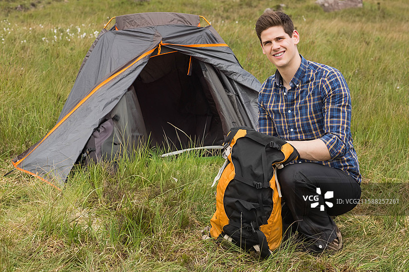男子跪在草地上的灰橙色圆顶帐篷旁，整理背包装备，留有空白空间。图片素材