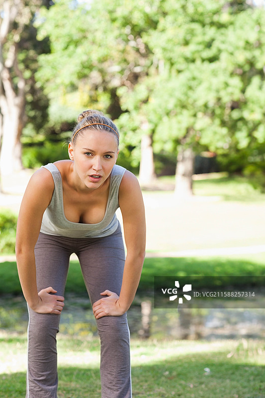 女性运动员在公园靠近倒影池塘处向前弯腰，喘息。图片素材