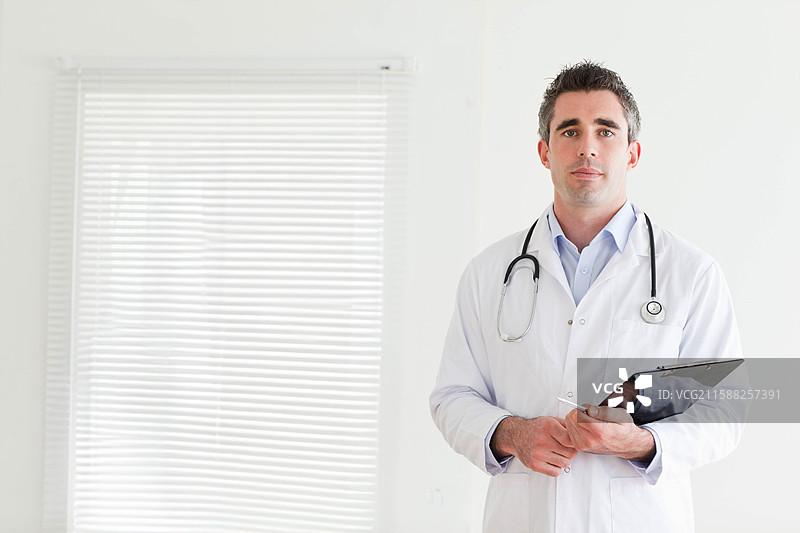 男医生站在诊所里，手持夹板和听诊器，靠近百叶窗，留有空白空间。图片素材