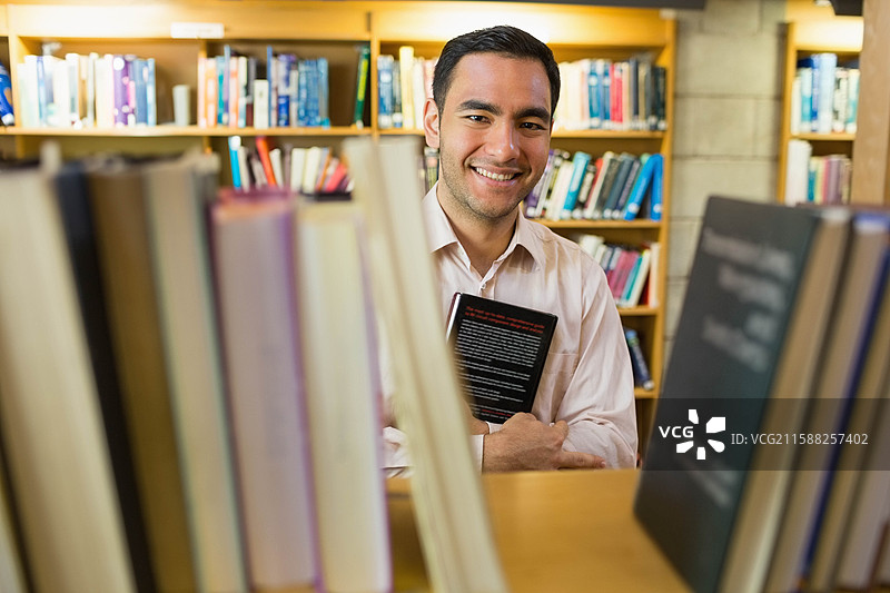 男学生站在图书馆的学习桌旁，手里拿着一本精装书，面带微笑，周围是书架。图片素材