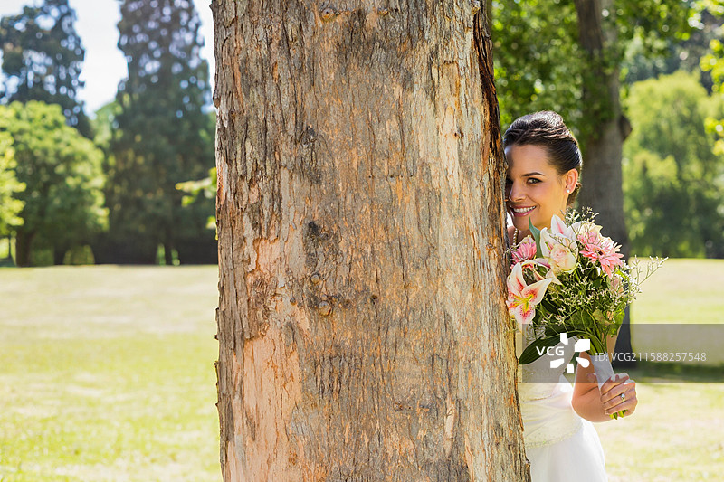 穿着婚纱的女人在公园的大树干后面探头，手里拿着花束，留有空白空间。图片素材