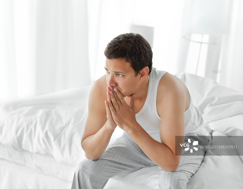 男人坐在床边，身体前倾，双手捂住嘴，卧室里铺着白色床单。图片素材