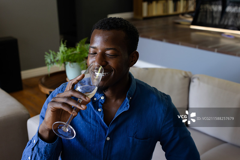 非裔美国男性在现代客厅的米色沙发上喝白葡萄酒。图片素材