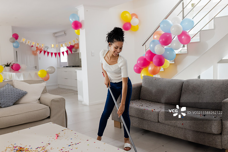 青春女孩在现代开放式客厅中用扫帚清扫五彩纸屑，周围有气球，留有空白空间图片素材