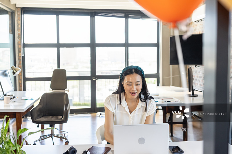 多元化女性在阳光明媚的开放式办公室桌子上使用笔记本电脑，旁边有智能手机和橙色气球。图片素材