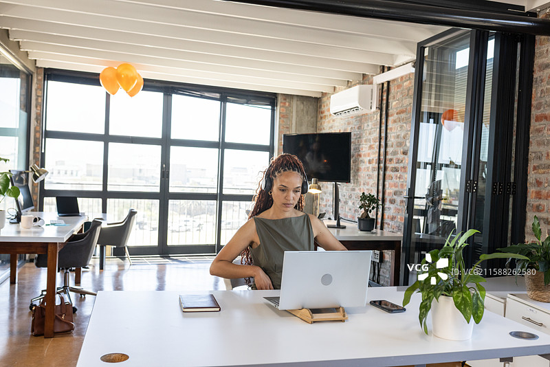 非裔美国女性在办公桌上使用笔记本电脑，旁边有笔记本、智能手机和耳机。图片素材
