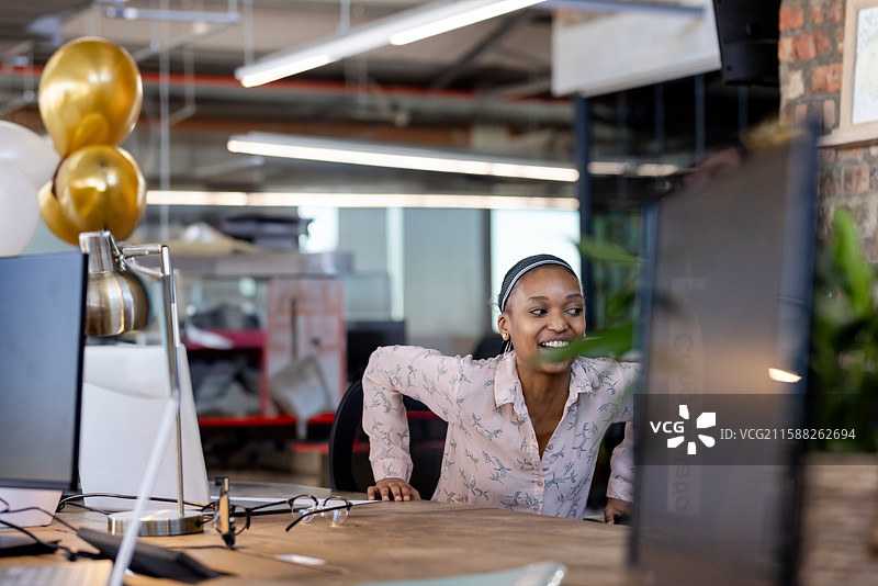 非裔美国女性在办公室的桌子前向前倾身，使用带有金色气球的电脑显示器。图片素材