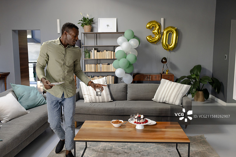 非裔美国男性在客厅的桌子上摆放平板电脑，旁边有生日蛋糕和气球。图片素材