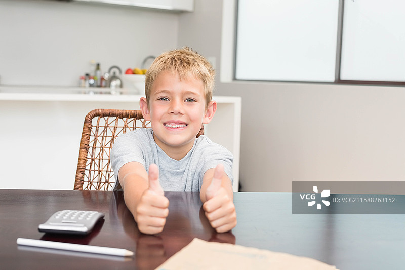 微笑的男孩坐在家里的深色木桌旁，正在用计算器和铅笔在纸上计算。图片素材