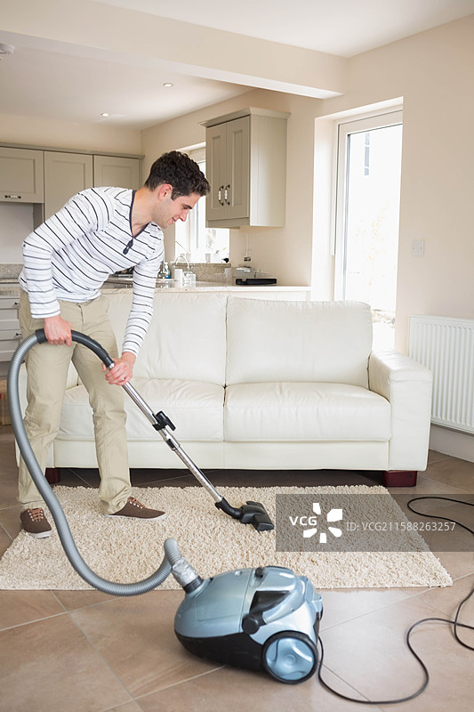男子在客厅的瓷砖地板上用吸尘器清洁区域地毯，靠近厨房橱柜。图片素材