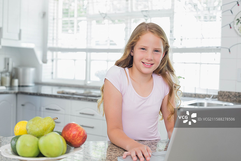 学龄女孩靠在厨房的花岗岩台面上，一边使用笔记本电脑，一边吃水果。图片素材