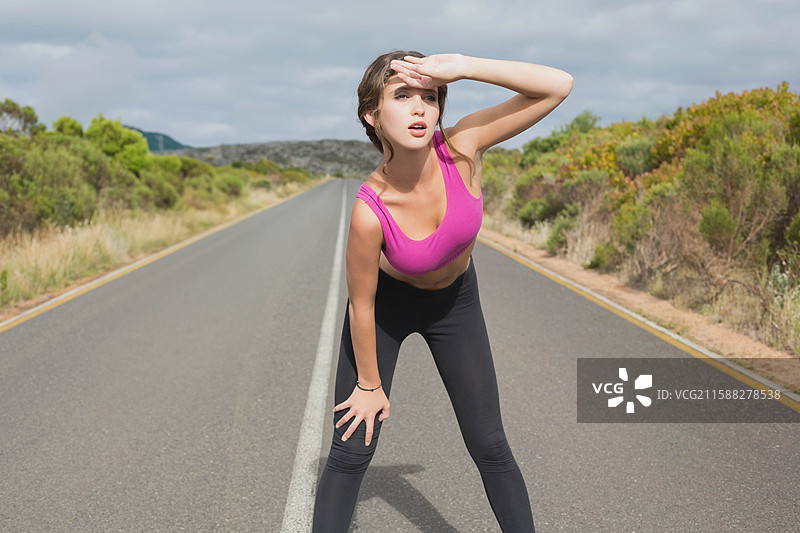 穿着粉色运动胸罩和黑色紧身裤的女性向前倾身，遮住眼睛，站在乡村铺砌的道路上。图片素材