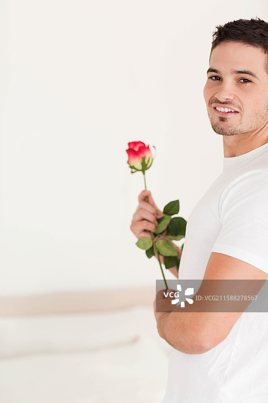 穿着简单白色T恤的男子站在明亮的卧室里，手中捧着红白玫瑰，留有空白空间。图片素材