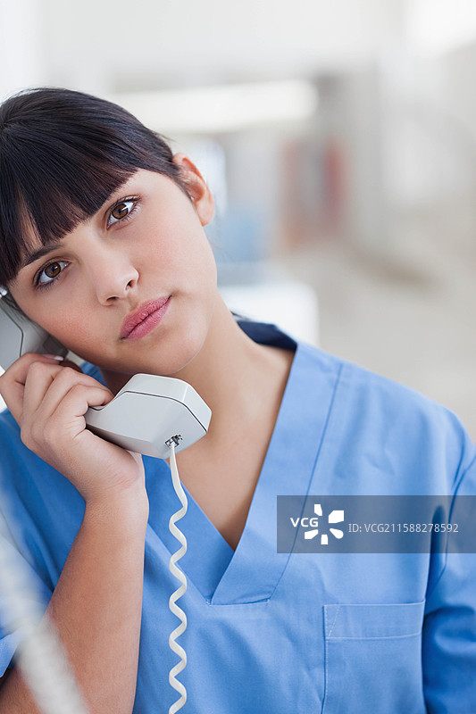 穿着蓝色护士服的女性护士站在医院走廊，手里拿着电话听筒。图片素材