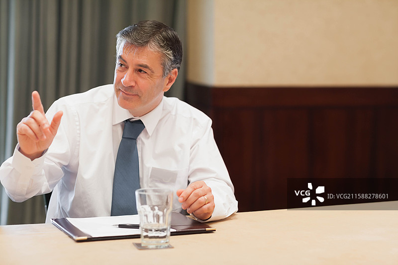 中年男子穿着西装，在会议室的桌子旁举起手指，桌上有文件夹、笔和水杯。图片素材