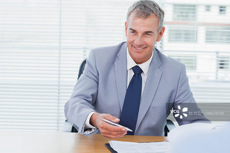 老年男子在办公室的木桌旁，窗帘旁边，手持钢笔，俯视一堆文件。图片素材