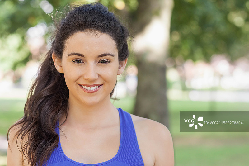 穿着明亮蓝色运动背心的女性在公园里微笑着直视镜头。图片素材