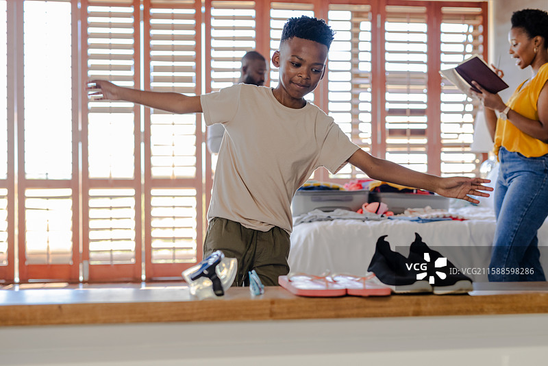 男孩在家中快乐地平衡在鞋子旁，父母则在整理衣物。图片素材