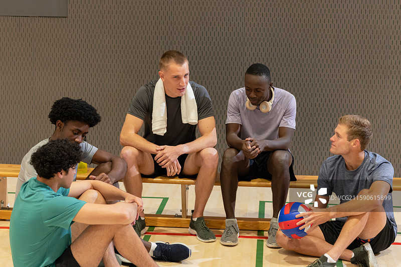 男性朋友们穿着运动服，坐在体育馆的长椅和地板上休息，手里拿着排球。图片素材