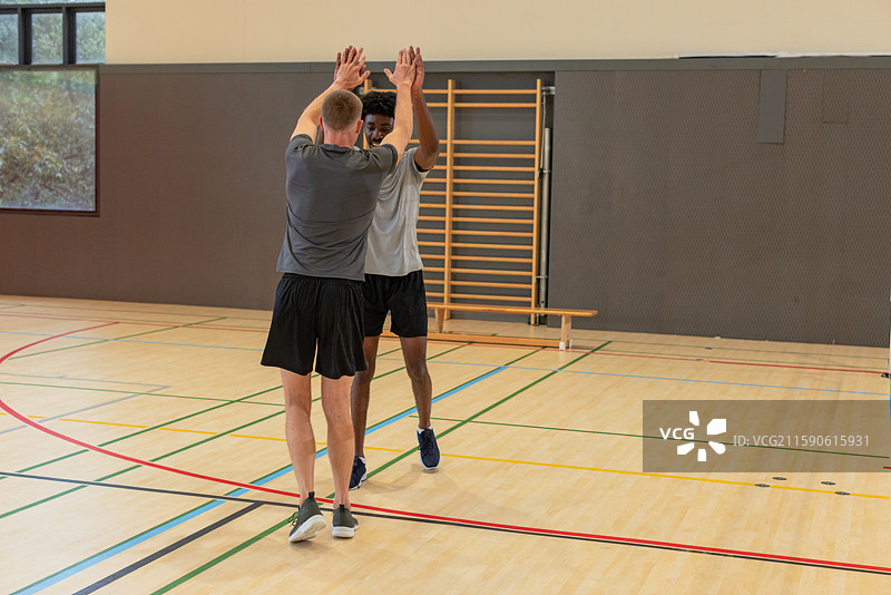 多样化的男性健身伙伴在攀爬架旁的体育馆中举起双臂，场地线条清晰可见图片素材