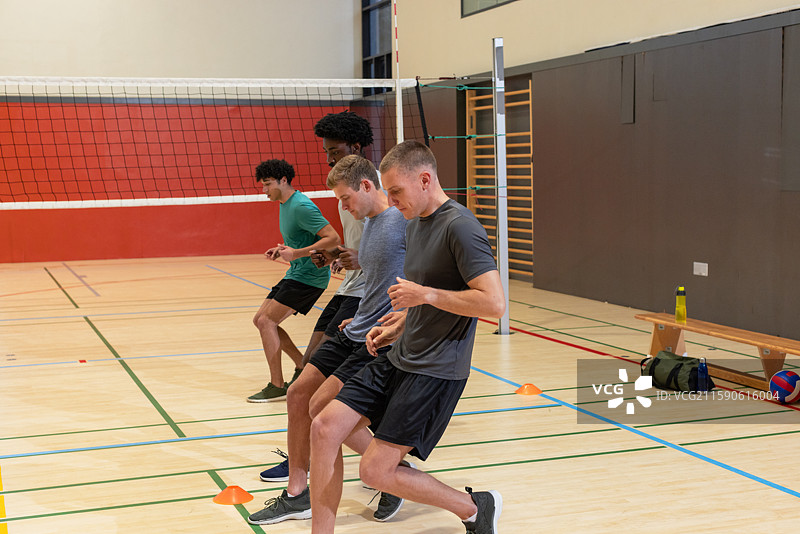多位不同种族的男性运动员在健身房练习脚步训练，旁边有排球网和橙色锥形标志。图片素材