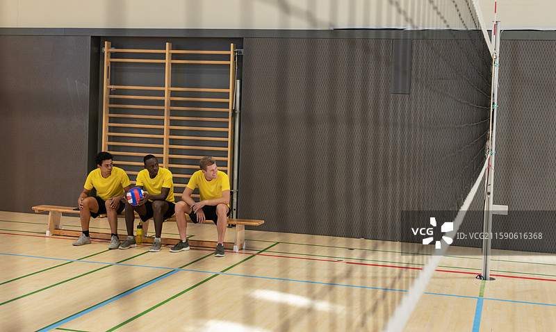 多元化的男性排球搭档坐在学校体育馆的长椅上，手里拿着球，靠近网，留有空白空间。图片素材