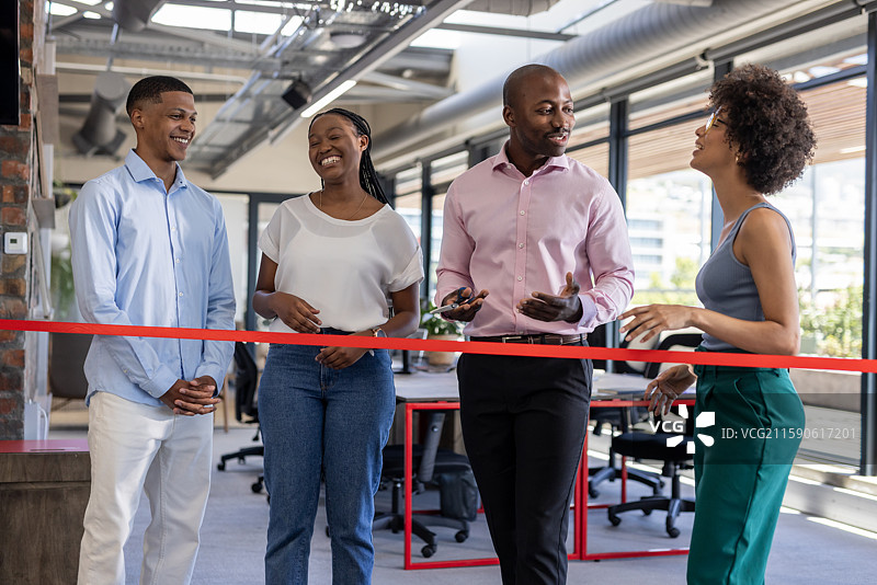 同事们在室内庆祝办公室开业，微笑着聊天，旁边有红色丝带。图片素材