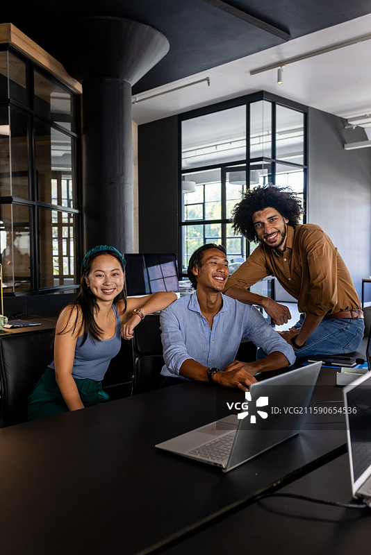 在办公室，来自不同背景的商业团队围绕笔记本电脑合作，自信地微笑。图片素材