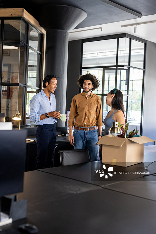 现代办公室的同事们在桌子旁聊天并微笑，旁边有一个未拆封的箱子。图片素材