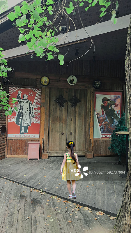 时光街角的童趣 穿黄裙的小女孩走向木楼，楼旁革命宣传画与葱郁绿叶相映，满是岁月与童真的交融。图片素材