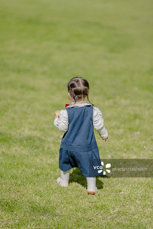 草坪上散步的穿蓝色背带裙女孩背身高角度视图图片素材