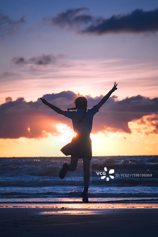 这张照片展现海边日落，少女剪影跳跃，双臂舒展，霞光漫天，海浪轻涌，满是自由欢快的气息。图片素材