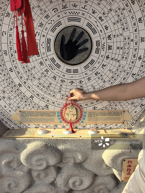 一只手握着带红绳的祈福挂饰，背景是刻有八卦、干支的罗盘，中心有黑色手掌印，充满传统民俗祈福氛围。图片素材