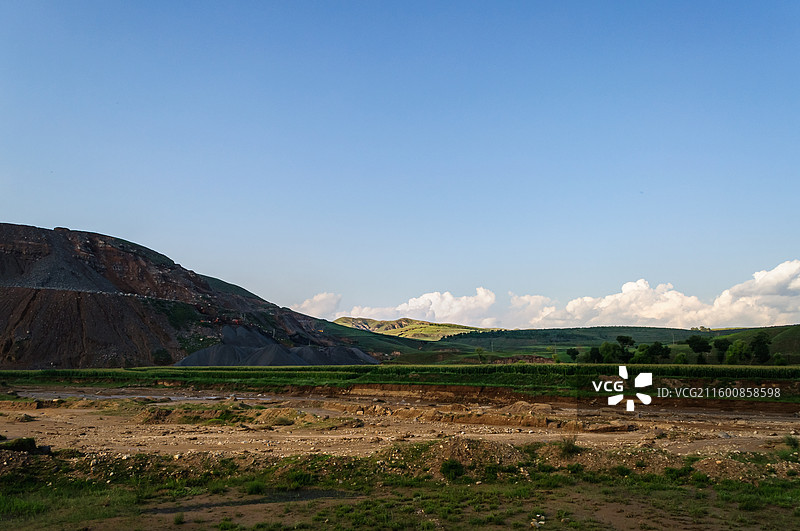 蓝天白云下开阔的山地田野景观，前景荒芜土地与中景绿意农田形成层次分明的构图图片素材