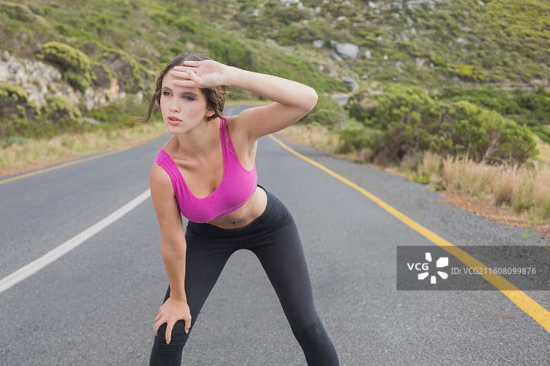 穿着粉色上衣和黑色打底裤的女士在山路上驻足，用手遮挡眼睛，路边有白色中心线图片素材