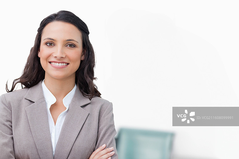 一位女士微笑着，双手交叉而站，身穿浅灰色西装外套，背景是办公室和一张绿松石色的椅子图片素材