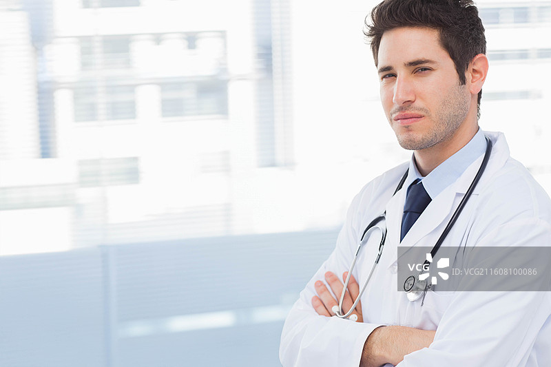 男医生双臂交叉站在诊所窗边，身穿白大褂，戴着听诊器，有复制空间图片素材