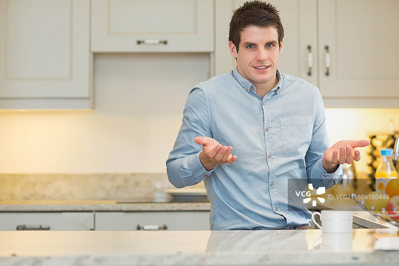 一名穿着蓝色纽扣衬衫的成年男性坐在厨房柜台上，双手掌心向上，示意动作，旁边放着一个马克杯图片素材