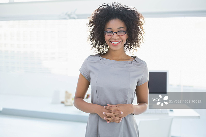 一位非裔美国女性站在办公室中心，脸上带着微笑，双手在办公桌旁交叠，戴着眼镜图片素材