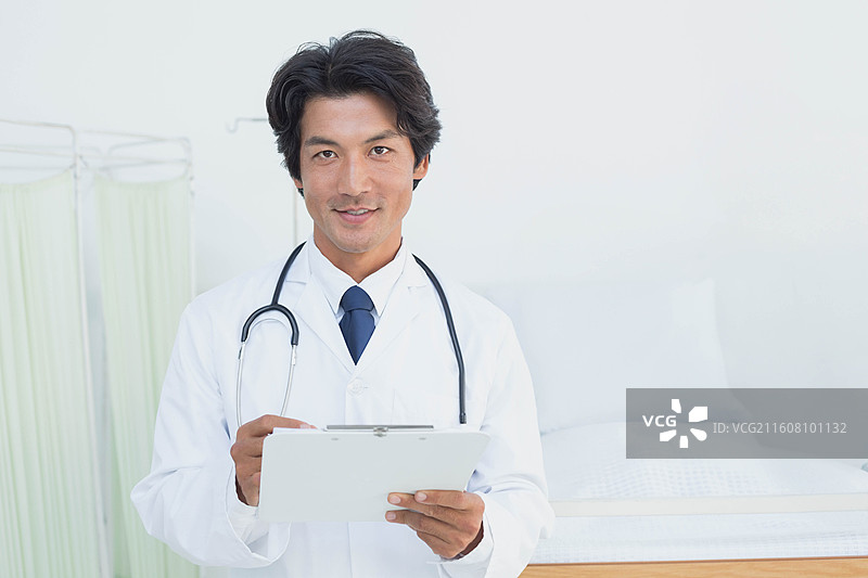 亚洲中年男性医生，穿着白大褂，戴着听诊器，在诊所用笔在病历夹上书写图片素材