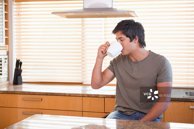 一名成年男子坐在厨房柜台前，用白色马克杯喝水，背景是石材台面，留有复制空间图片素材