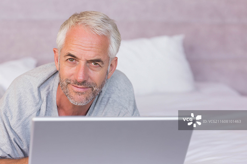 一位穿着灰色T恤的成熟男子，在卧室里用银色笔记本电脑，肘部支撑着身体躺在床上图片素材
