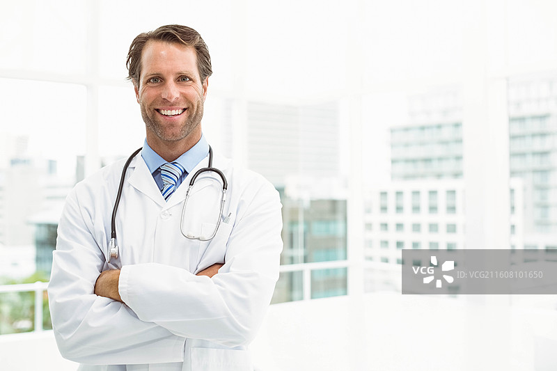 中年男医生身穿白大褂，佩戴听诊器，站在诊室靠窗处，旁边有空白空间可供编辑图片素材