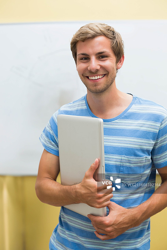 一个穿着蓝色条纹T恤的男人站在教室里，微笑着，手里拿着笔记本电脑，旁边是白板图片素材
