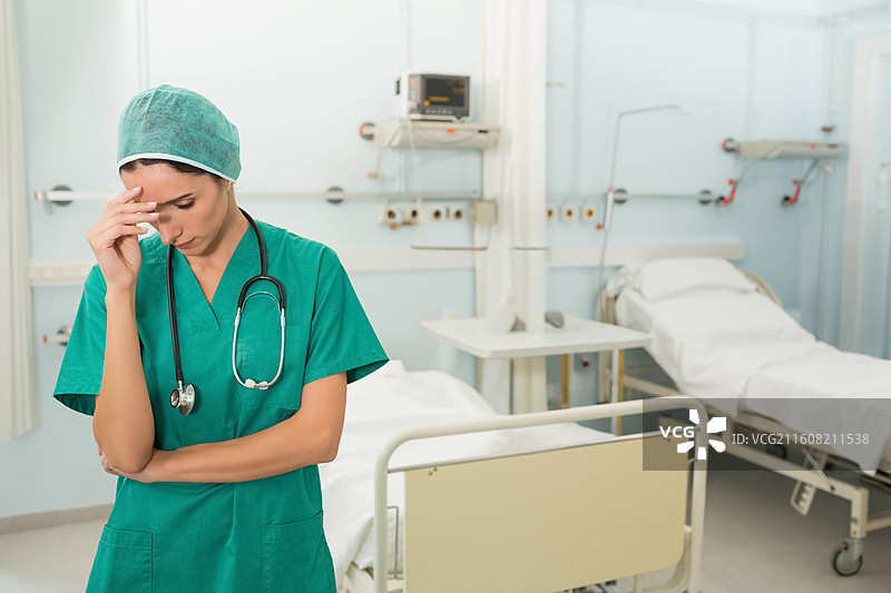 一名女护士穿着手术服，戴着听诊器，站在病房的床边，托着额头，留有复制空间图片素材
