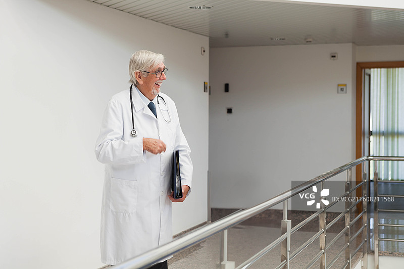 一位身穿白大褂、佩戴听诊器的资深男性医生，手持病历夹，站在医院走廊里交谈。图片素材
