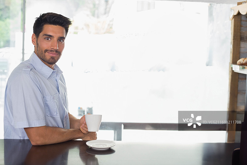 穿着蓝色衬衫的男人坐在木制咖啡馆柜台前，手里拿着带碟子的白马克杯，留有复制空间图片素材