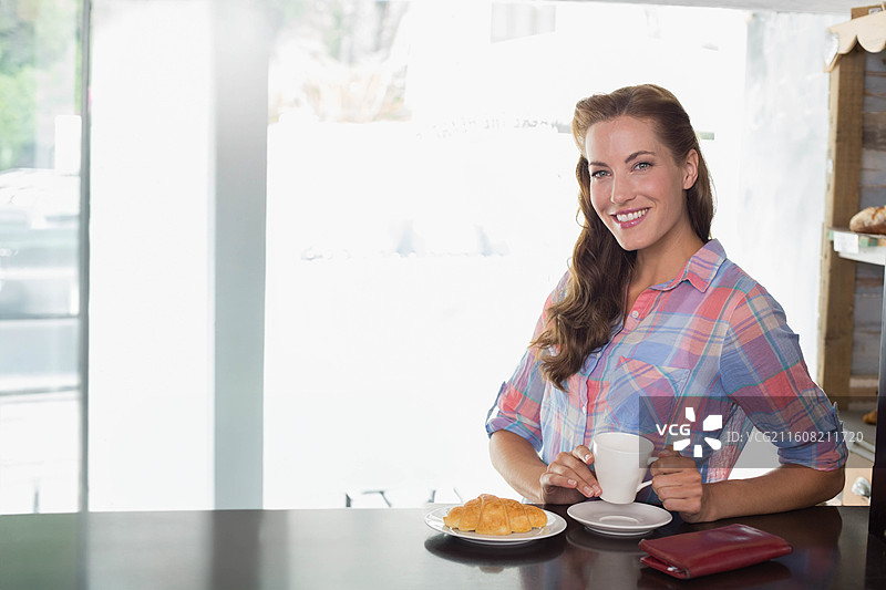 穿着淡色衬衫的中年女性，坐在咖啡馆吧台前，手持杯子和牛角面包，有空白区域可供添加文字图片素材
