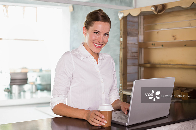 三十多岁的女性站在咖啡馆柜台前，身穿白衬衫，手持外卖杯，并使用笔记本电脑。图片素材